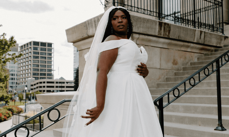 Plus-size bride wearing an off-the-shoulder A-line wedding dress and veil, standing on outdoor steps.