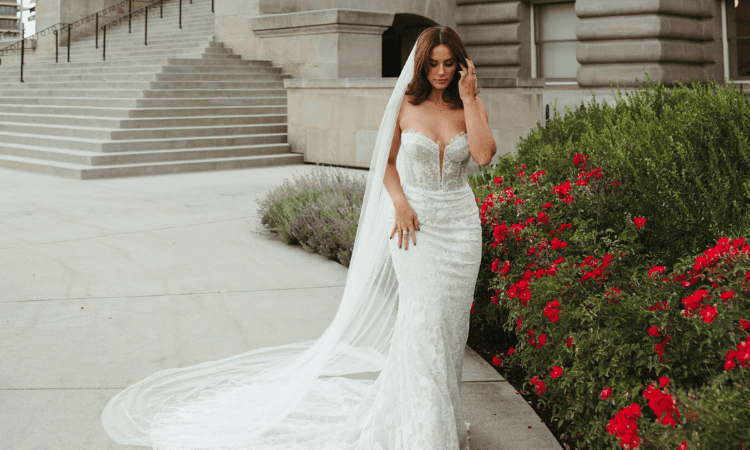 Bride wearing a fitted lace wedding dress with a long flowing veil and train, standing beside a garden of red flowers.