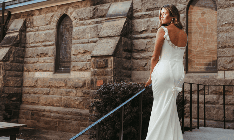 Bride wearing a fitted satin wedding dress with a low back and bow detail, standing beside a historic stone building.