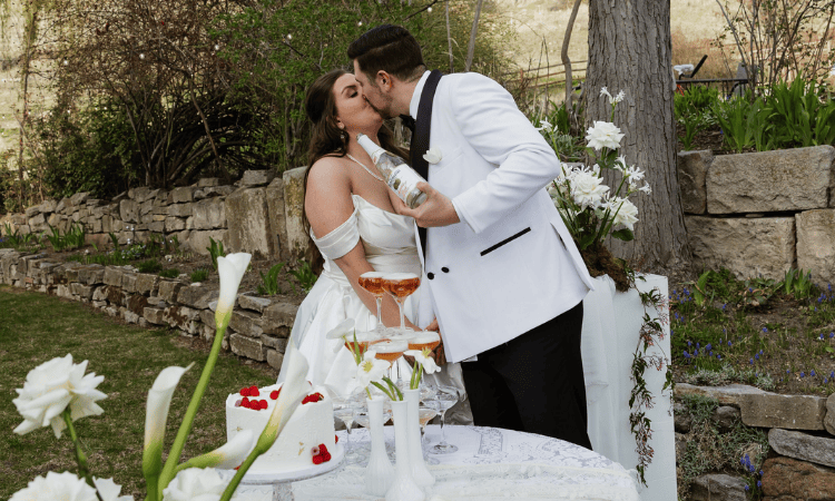 Newlywed couple kissing beside an outdoor wedding table with cake, flowers, and champagne.