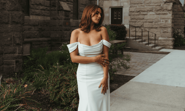 Bride wearing a fitted off-the-shoulder wedding dress with draped detailing and short sleeves, standing outdoors in front of a stone building.