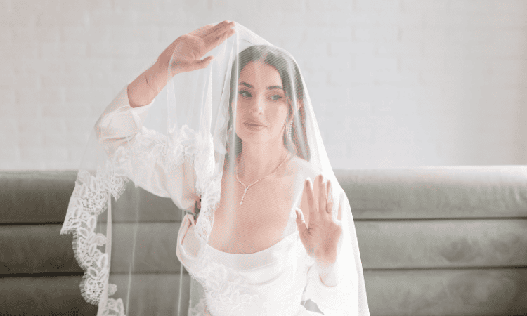Bride wearing a wedding dress and veil, gently lifting the veil while posing indoors.