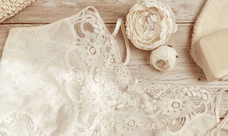 Close-up of white lace wedding dress fabric with floral detailing and bridal accessories on a wooden surface.