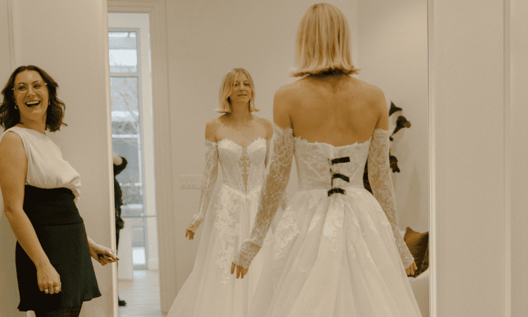 Bride viewing the back of her lace wedding dress in a mirror while a bridal consultant stands nearby.