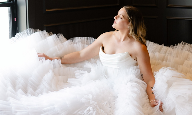 Bride wearing a dramatic tulle ball gown wedding dress sitting indoors by a window.