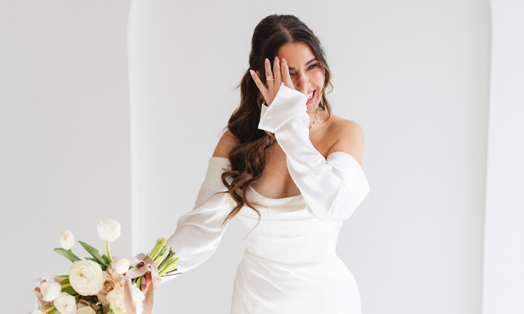 Bride wearing an off-the-shoulder satin wedding dress holding a bouquet and smiling.