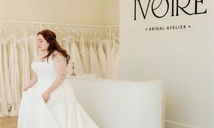 Plus-size bride wearing a white wedding dress in a bridal boutique with wedding gowns displayed behind her.
