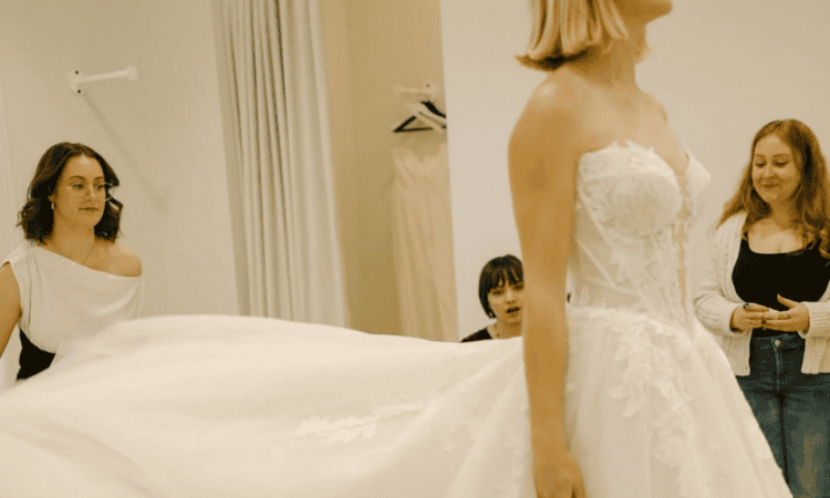 Bride wearing a strapless lace wedding dress while two women look on during a bridal dress fitting.