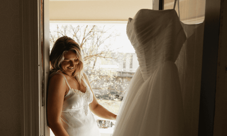 Bride smiling while looking at a wedding dress displayed by a window, with a second white gown hanging in the foreground.