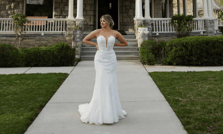 Bride wearing a fitted lace wedding dress with a plunging neckline and train, standing outside a stone mansion.