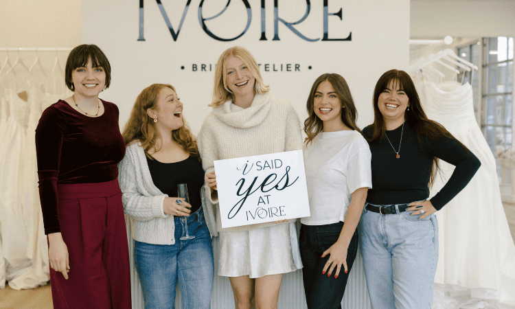 Bride and her friends posing with a sign reading “I said yes at Ivoire,” surrounded by wedding dresses.