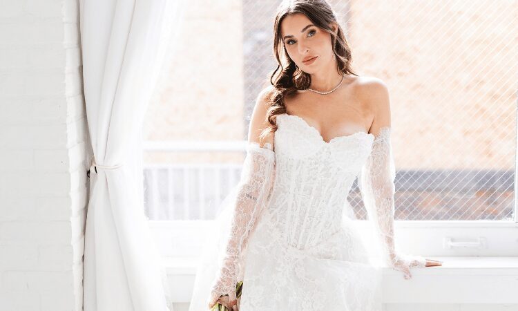 Bride wearing an off-the-shoulder lace wedding dress with long sleeves, standing by a window.