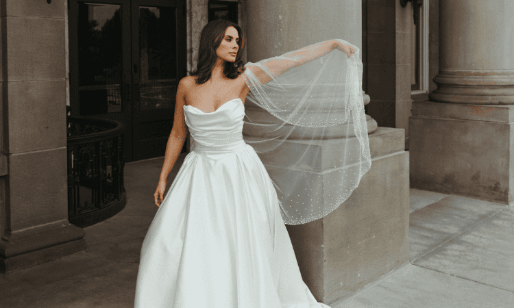 A bride in an elegant A-line wedding dress with a strapless draped bodice stands outside a grand building, holding a sheer embellished veil.